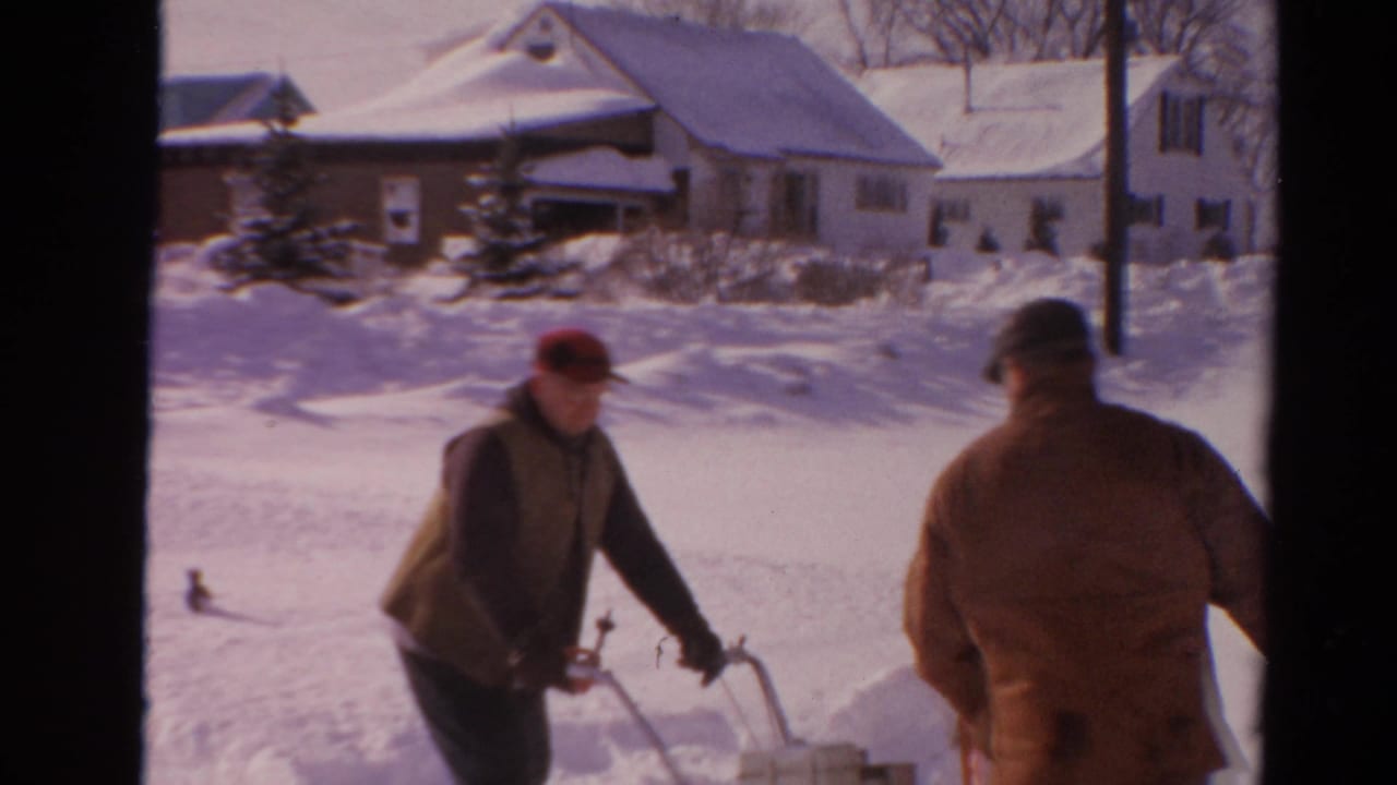 Two men stand near a snow blower on a snowy street with houses behind them.