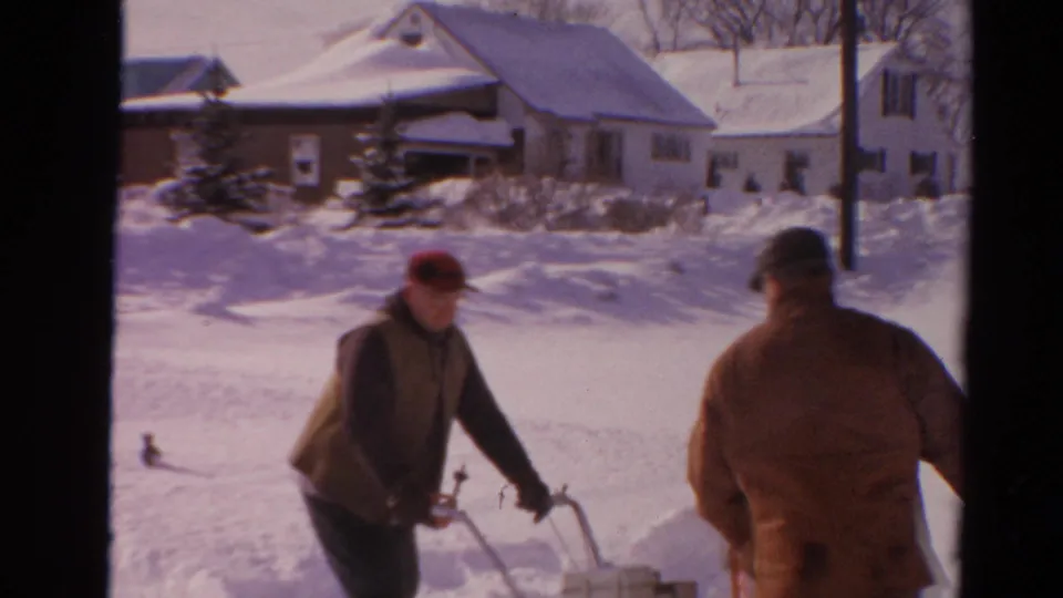 1954 Toledo frame with one man at a snow blower and another man approaching with a shovel on a snowy residential street.