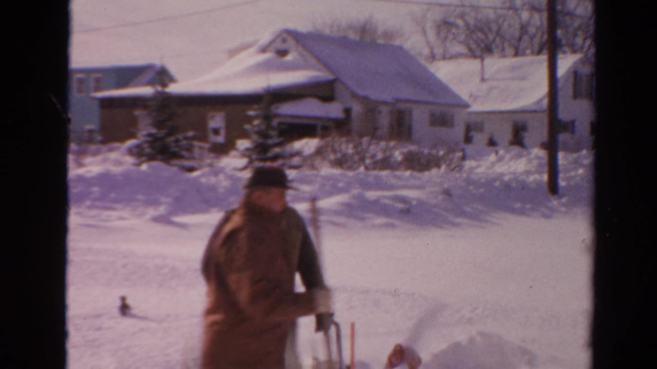 1954 Toledo home-movie frame showing a man shoveling near a snow blower in front of snow-covered neighborhood houses.