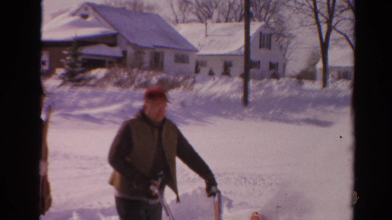 1954 Toledo frame showing the snow blower operator near a high snowbank, with houses and bare winter trees in the background.