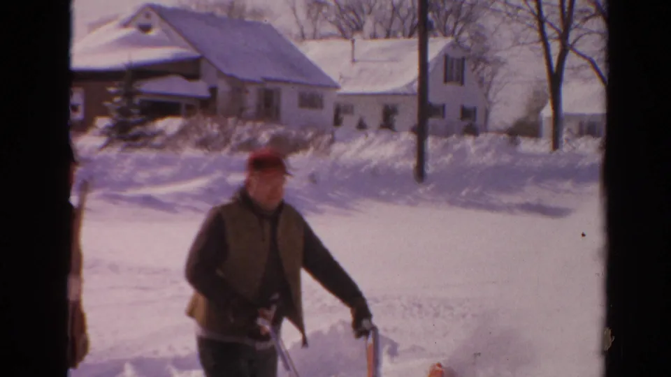 1954 Toledo frame showing the snow blower operator near a high snowbank, with houses and bare winter trees in the background.