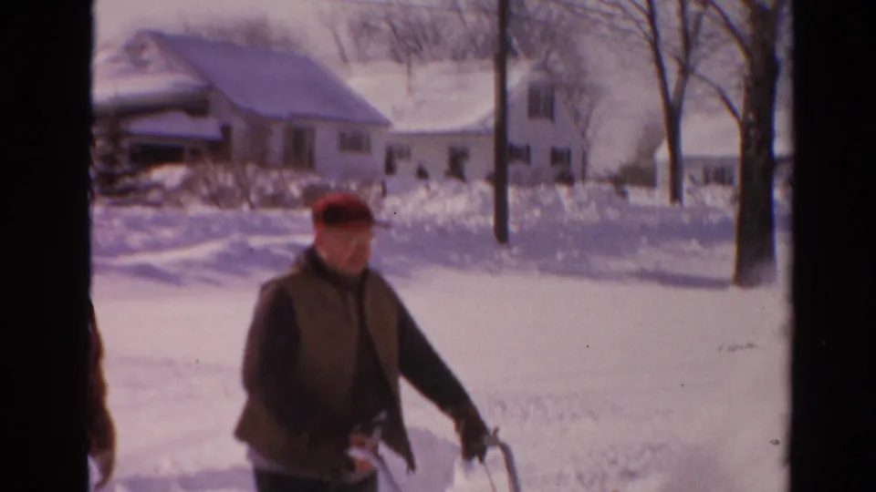 In 1954 Toledo, a man in a red cap and brown vest clears snow near detached houses on a bright winter day.