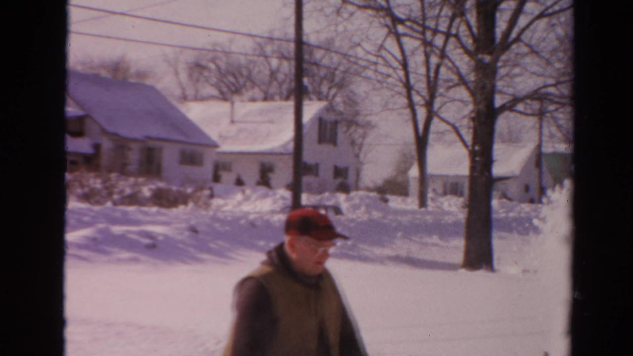 A soft 8mm frame shows the man partly bent while clearing snow, with Toledo residential houses and leafless trees behind him.