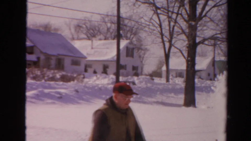 A soft 8mm frame shows the man partly bent while clearing snow, with Toledo residential houses and leafless trees behind him.