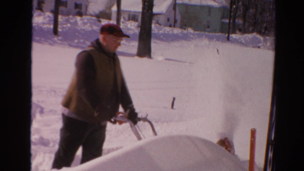 The man in a red cap walks past a snowbank as the snow blower throws snow beside a bright 1954 Toledo residential street.