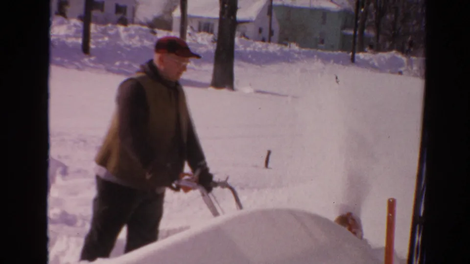 The man in a red cap walks past a snowbank as the snow blower throws snow beside a bright 1954 Toledo residential street.