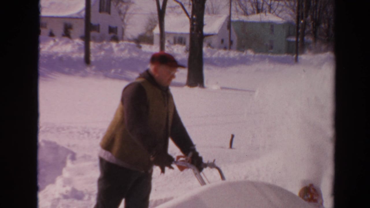 Man in a red cap uses a snow blower beside a snowy driveway.