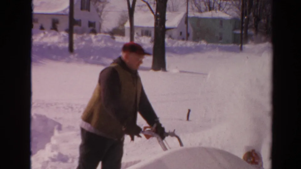 In Toledo, Ohio, 1954 home-movie footage, a man in a red cap operates a snow blower near piled snow and houses.