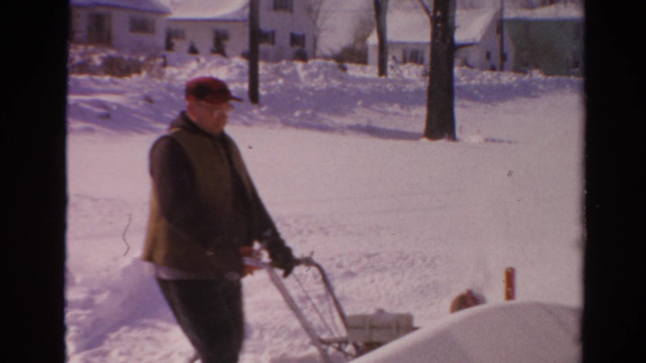 Man pushes a snow blower across a snow-covered residential yard or driveway.