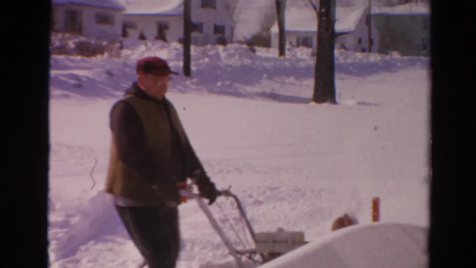 A man in winter clothing pushes a walk-behind snow blower in a snowy Toledo residential setting from 1954 footage.