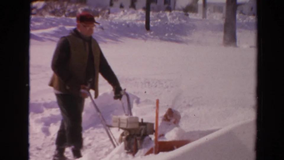 A man operating a snow blower pauses near a shovel and high snowbanks in 1954 Toledo home-movie footage.