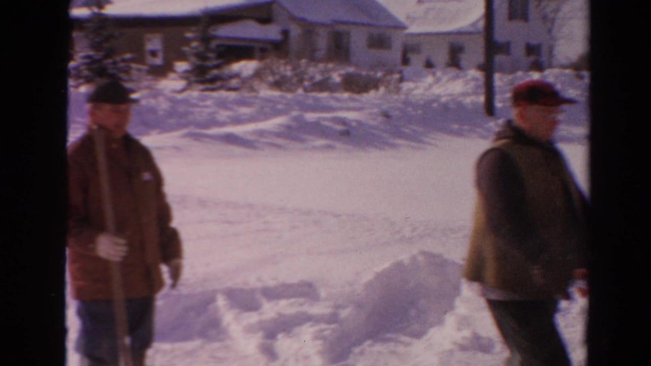 Two adult men appear in a snowy Toledo neighborhood, with one holding a shovel as the snow blower operator walks past.