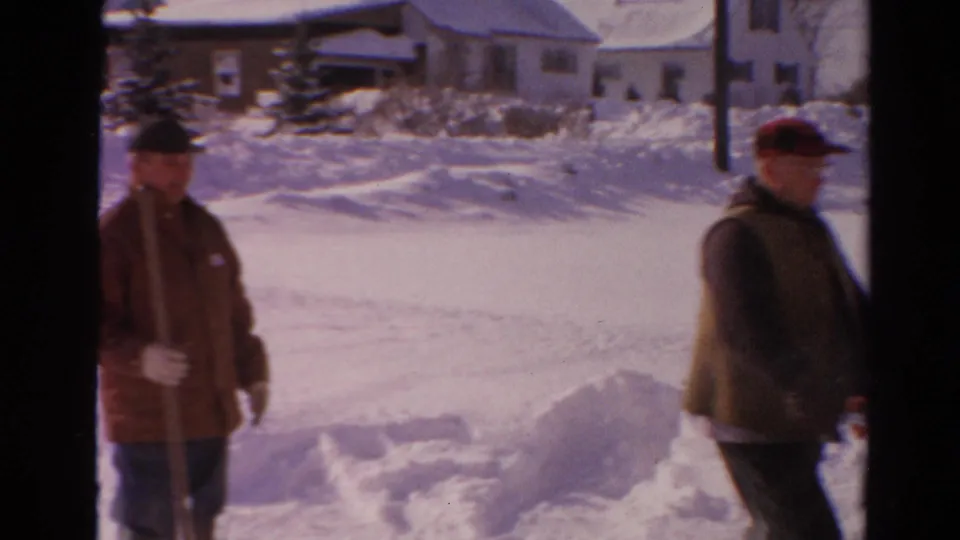 Two adult men appear in a snowy Toledo neighborhood, with one holding a shovel as the snow blower operator walks past.