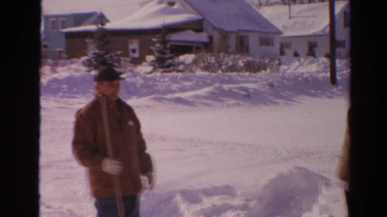 Man in a brown coat stands in deep snow near suburban houses.