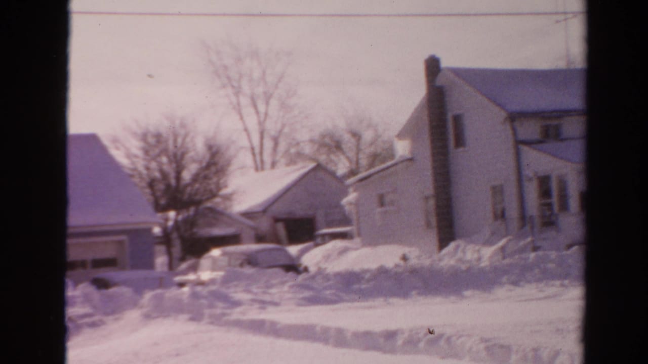 A snowy Toledo neighborhood view includes residential houses, a garage, partly buried parked cars, and bare winter trees.