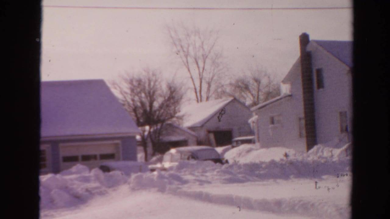Snow-covered suburban houses with a partly buried car near a driveway.