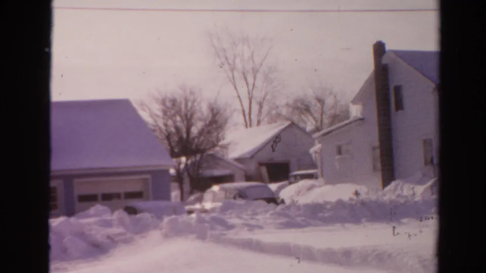 1954 Toledo home-movie frame showing deep snow around residential houses and a partly buried mid-century car.