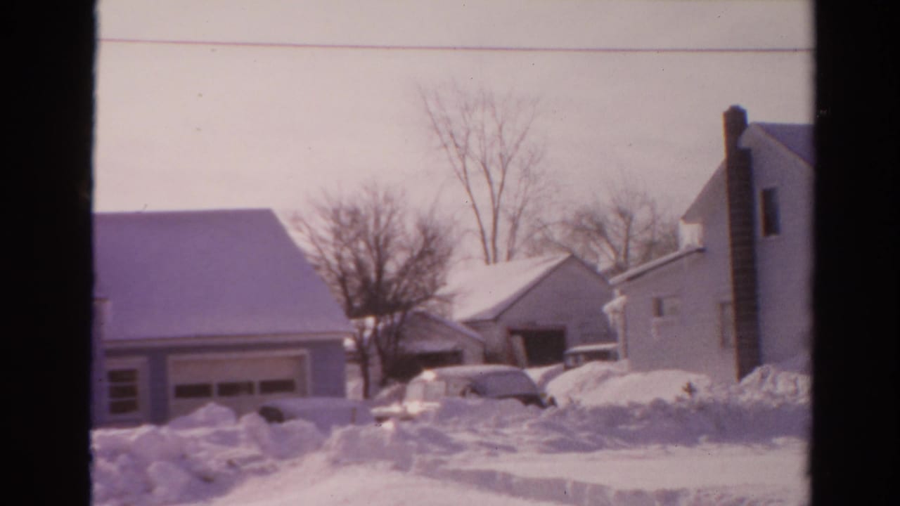 Residential street scene with snowbanks, houses, trees, and a car near a driveway.
