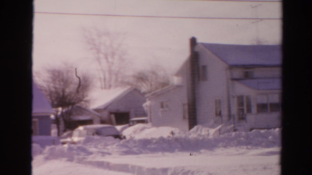 Soft 8mm frame from Toledo in 1954 showing a snowbound residential driveway, house fronts, and a partly visible car.