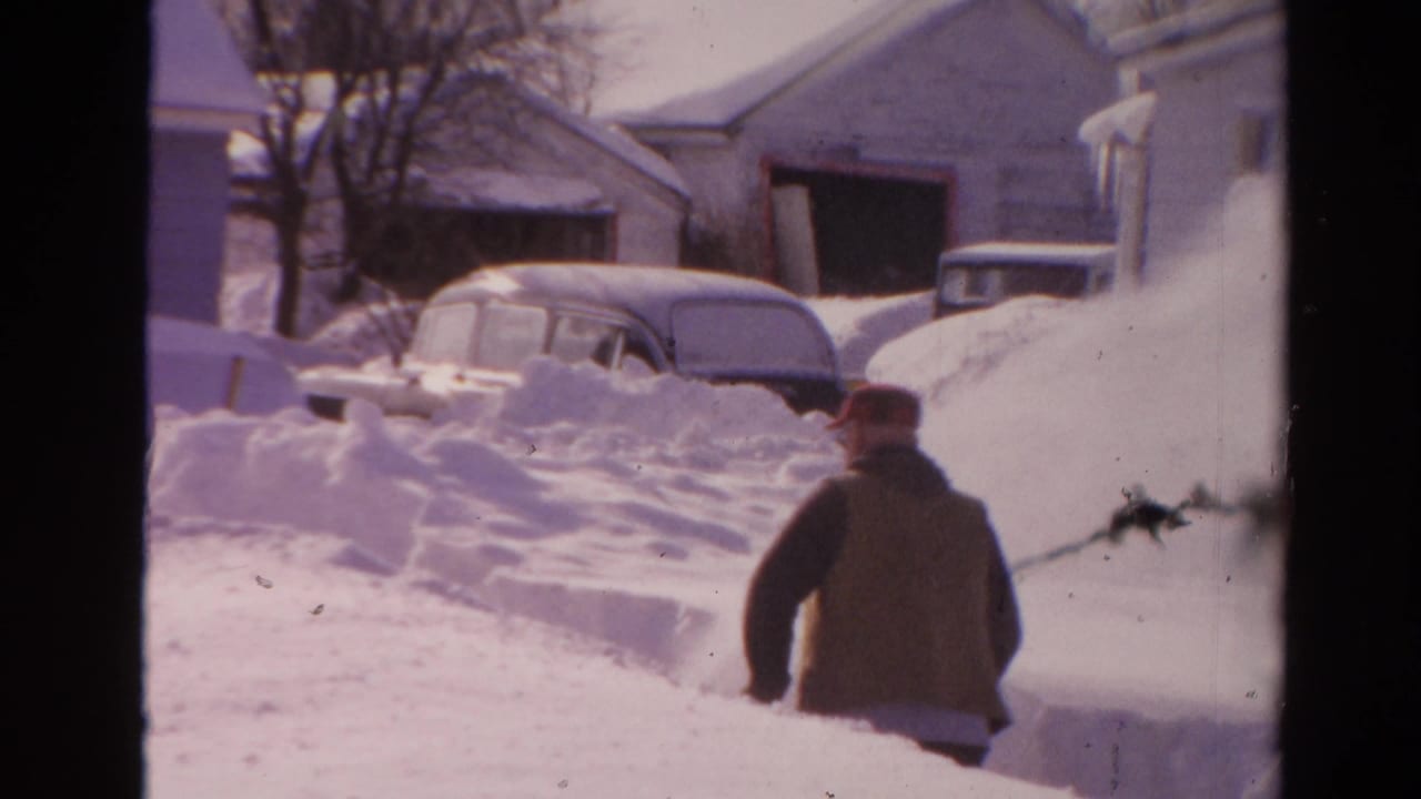 Man in a red cap walks through deep snow near houses and buried cars.