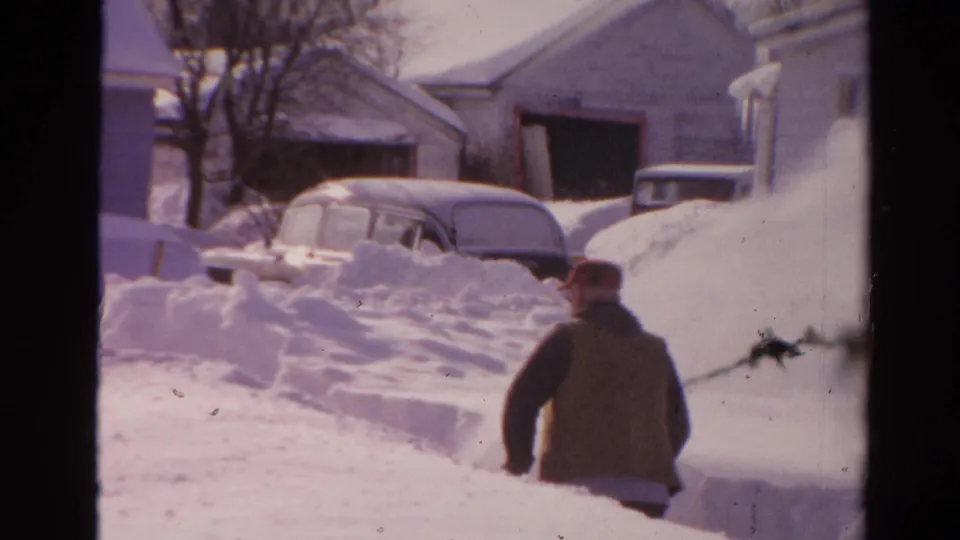 1954 Toledo home-movie frame showing a bundled man in a red cap amid deep snow, with houses and vintage cars in the background.