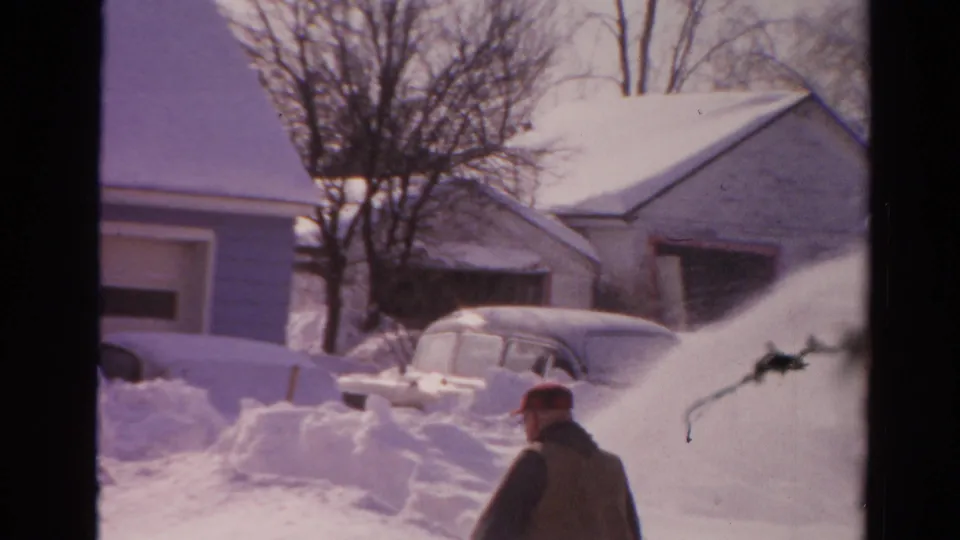 1954 Toledo winter frame with snow being thrown or blown across the view while a man works near snowbanks, houses, and older cars.