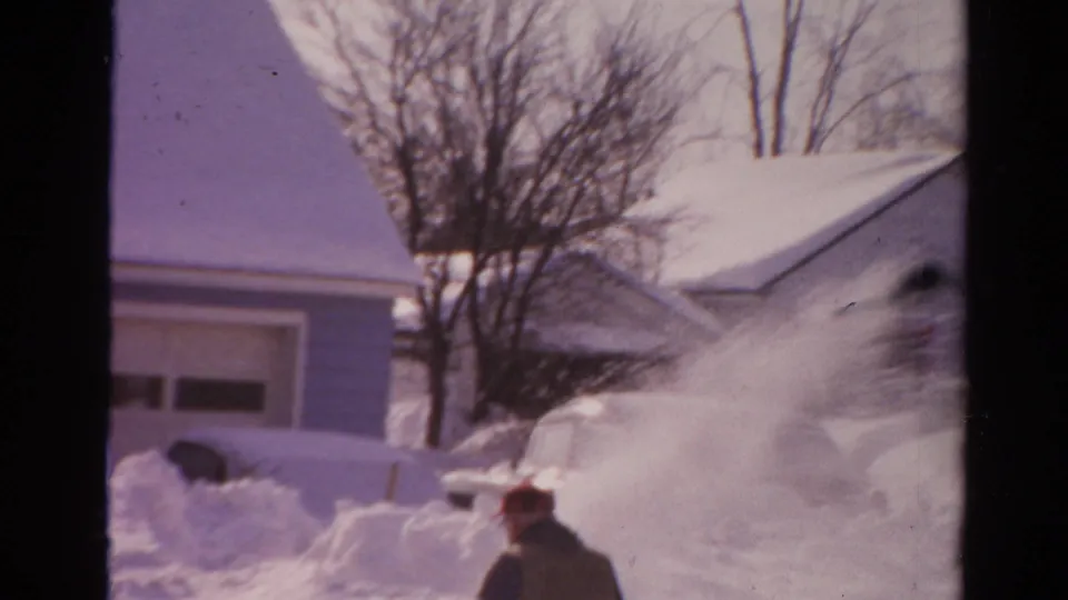 Home-movie frame from 1954 Toledo showing the man partly turned away as snow spray crosses a snowy residential scene.