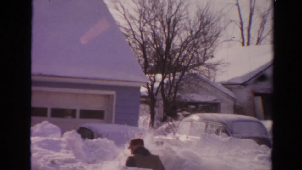 1954 Toledo frame showing a bundled man clearing heavy snow beside a partially buried car, with houses and bare trees behind him.