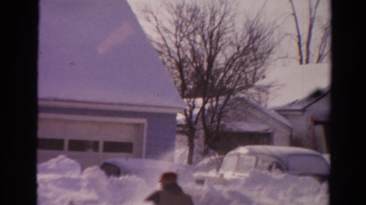 Man in a red cap works in deep snow near a garage and parked car.