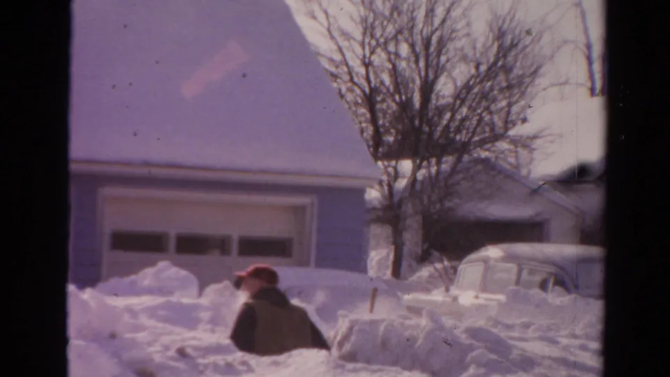 1954 Toledo winter frame with a man in a red cap partly surrounded by snowbanks near a garage and older parked car.