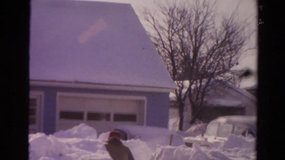 1954 Toledo home-movie frame showing the man low in heavy snow, with a garage, bare tree, and snow-covered car behind him.