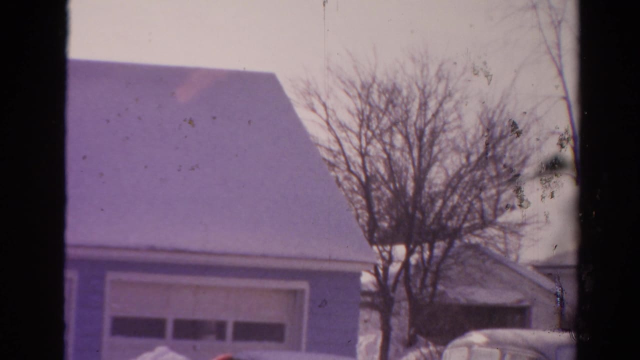 1954 Toledo home-movie frame showing a snow-covered suburban house, garage doors, bare trees, and deep snowbanks.