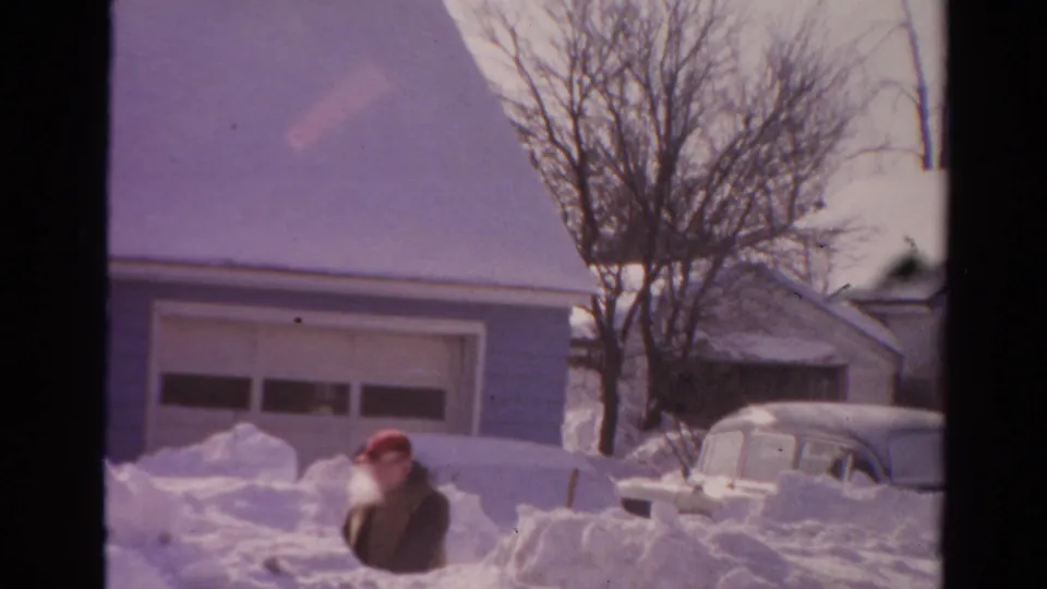 1954 Toledo frame with a man in a red cap operating near deep snow outside a suburban house and garage.