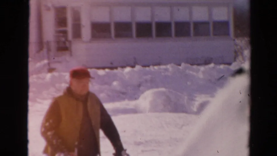 1954 Toledo frame showing a man in winter clothing beside a snowblower, with a snow-covered house and yard behind him.