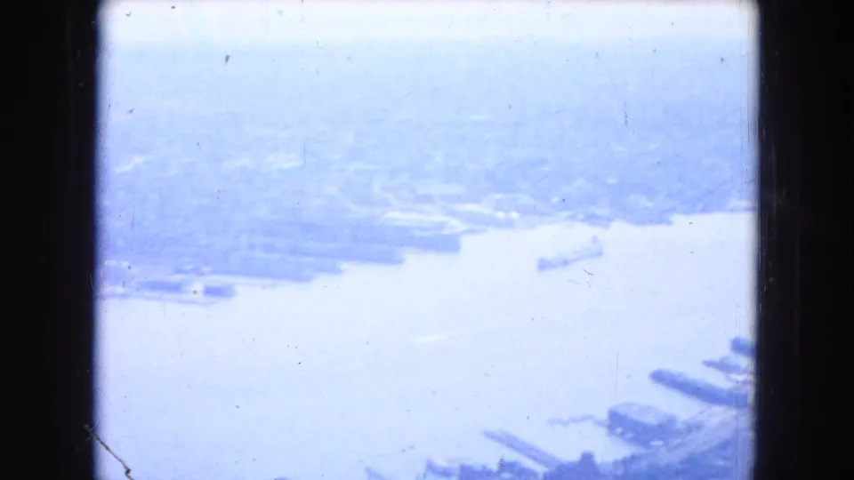 1954 New York aerial view with a broad river, hazy waterfront, and dark 8mm frame edges under cloudy light.