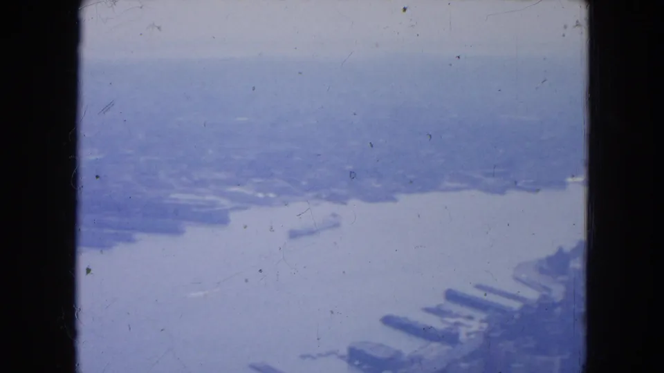 1954 New York aerial frame showing a river, shoreline, piers, and distant urban forms softened by haze and film wear.