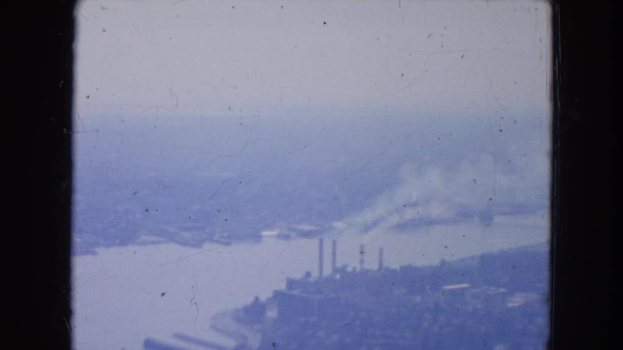 Hazy aerial view of a river, industrial waterfront, smokestacks, and distant shoreline.