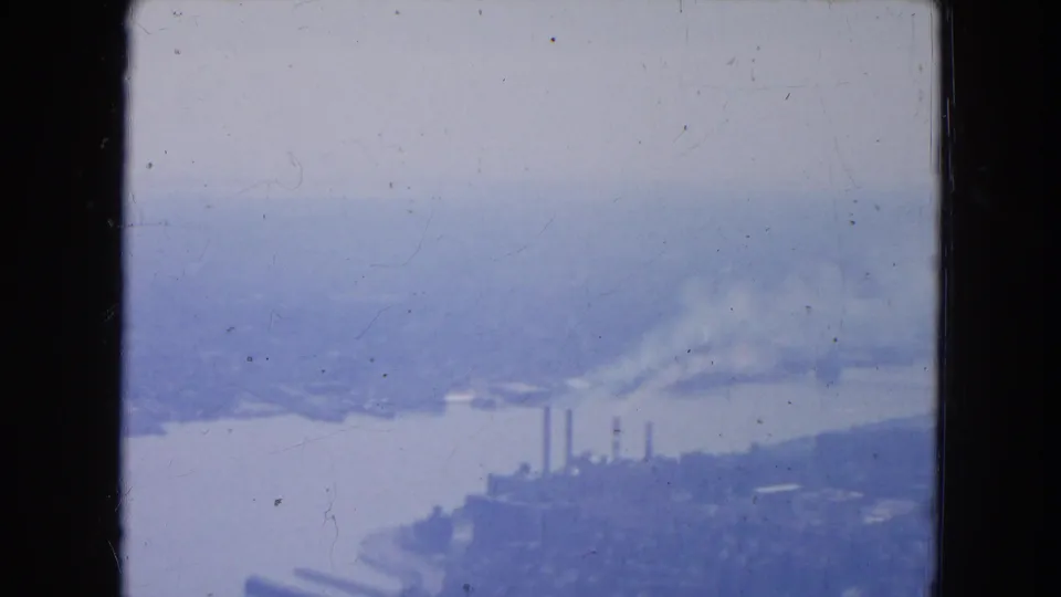 Blue-tinted 1954 New York City aerial frame showing a broad river, waterfront industry, smokestacks, and hazy distant land.