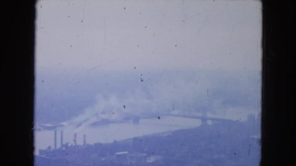 1954 New York City aerial view with a river crossing the frame, smoke over industrial buildings, and a soft hazy skyline.