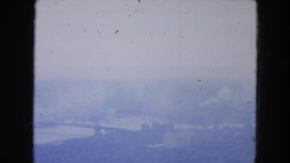 Soft, overexposed 8mm aerial frame from New York City showing a pale river view, low shoreline, and industrial forms in haze.