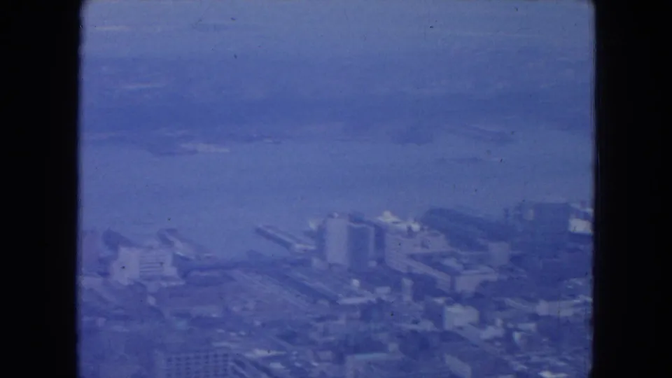 Blue-tinted 1954 New York City aerial frame showing water, shoreline buildings, possible piers, and dense waterfront blocks.