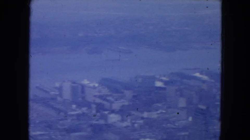 1954 New York City aerial view with waterfront buildings, broad river, cloudy sky, blue tint, and visible 8mm frame borders.