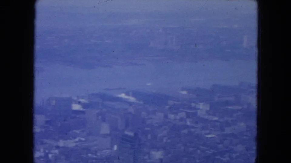 Blue-tinted 1954 New York City aerial frame showing a river, waterfront blocks, tall buildings, and cloudy haze.