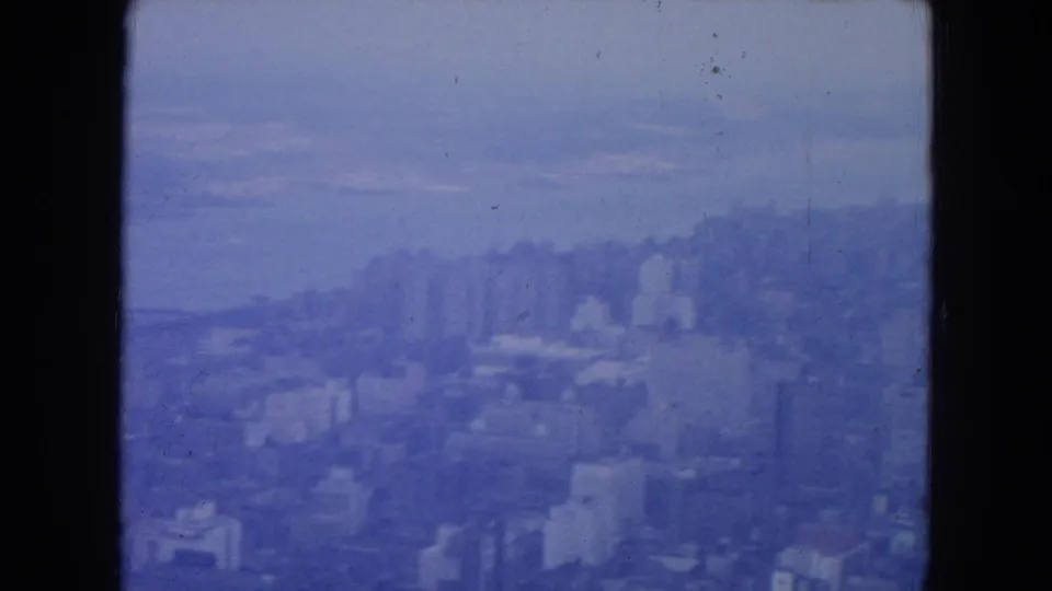 Blue-faded 8mm frame of New York City in 1954, showing clustered buildings and a hazy waterfront from above.