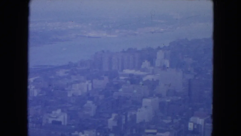 1954 high-vantage New York City view with mid-rise blocks in the foreground and a pale waterfront beyond.
