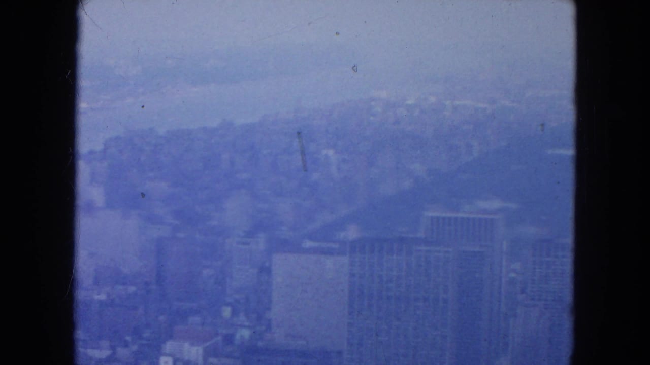 Hazy skyline seen through dark vertical window edges with mid-century buildings in blue tint.