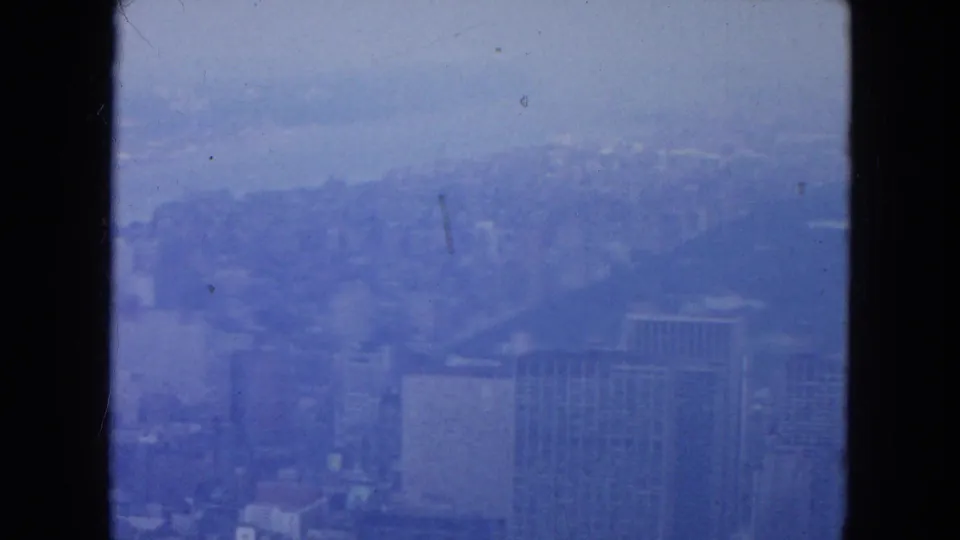 1954 New York City skyline appears through dark window framing, with high-rise buildings softened by haze and blue 8mm color cast.