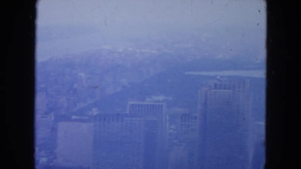 A hazy 1954 New York City skyline view shows tall buildings and a distant waterway, partially obscured by soft atmospheric haze.