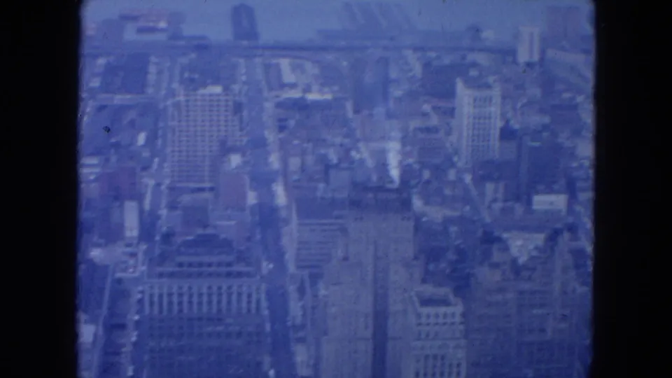 A downward 1954 New York City view shows dense streets, rooftops, tall buildings, and distant water through blue-tinted 8mm haze.