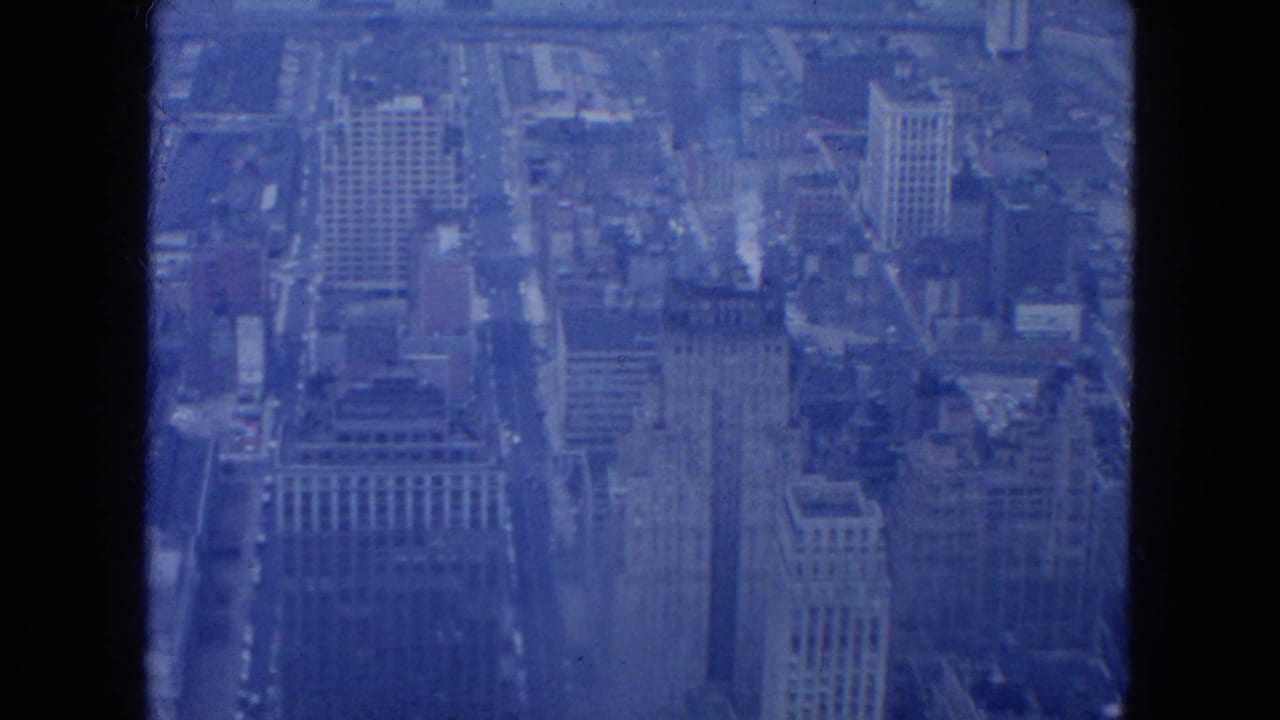 1954 New York City aerial frame showing tall commercial buildings and a traffic-lined avenue in soft blue 8mm color.